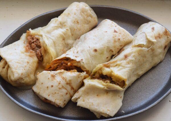 Three GRAB AND GO 4 OZ. New Mexico Burritos from two 2-packs, each with different fillings including meat and vegetables, are arranged on a round, dark plate.