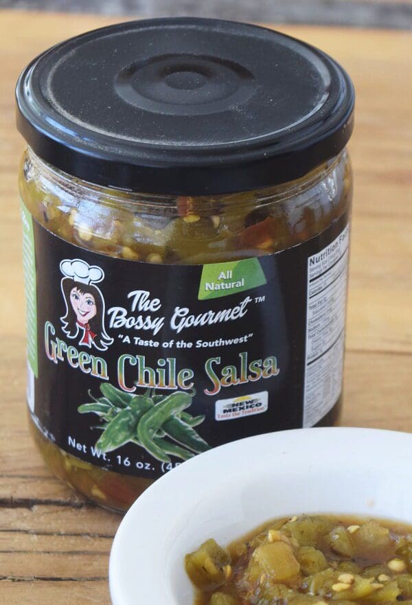 A jar of Hatch Green Chile Salsa, medium-hot flavor, on a wooden surface, with a small bowl of salsa in front. The label indicates it is all-natural and 16 ounces.