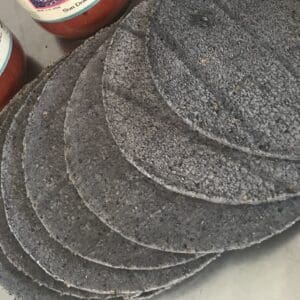 Two packages of blue corn tortillas, each containing 12 tortillas, are arranged next to jars of sundried tomato sauce on the countertop.