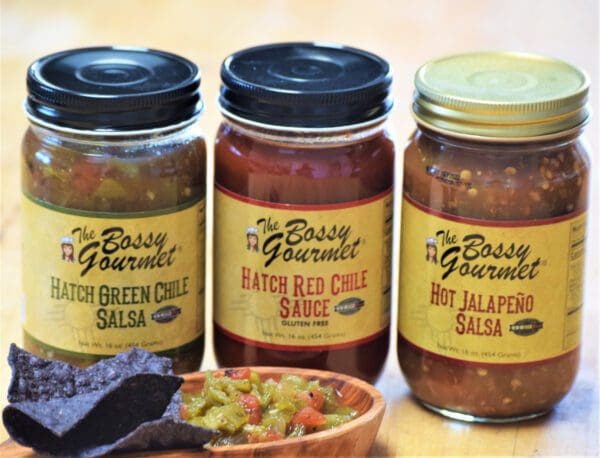 Image of three jars of Authentic New Mexico Salsas - Choice of Flavors: Hatch Green Chile Salsa, Hatch Red Chile Sauce, and Hot Jalapeño Salsa. Blue corn tortilla chips and a salsa dip are placed in front of the jars.