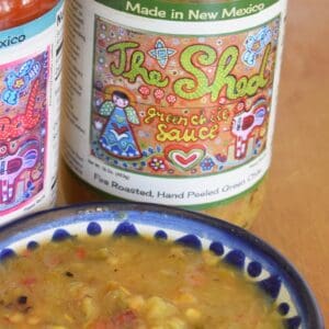 A 16 oz. jar of GREEN CHILE SAUCE FROM THE SHED with a bowl of the sauce in front. The label on the jar reads "Made in New Mexico" and features colorful artwork, while another jar with a red lid is partially visible beside it.