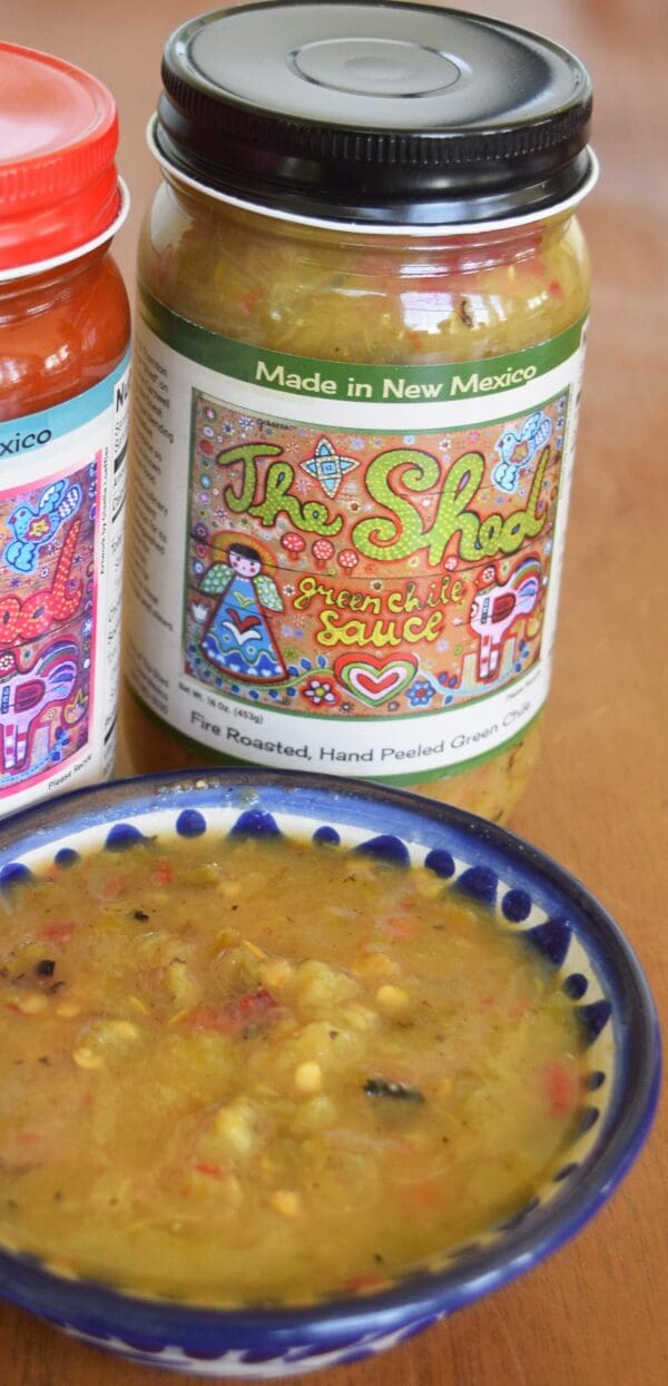 A 16 oz. jar of GREEN CHILE SAUCE FROM THE SHED with a bowl of the sauce in front. The label on the jar reads "Made in New Mexico" and features colorful artwork, while another jar with a red lid is partially visible beside it.