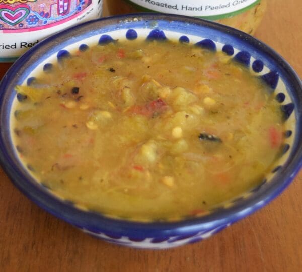 A bowl of green chile sauce from The Shed with visible seeds and veggie chunks in a blue and white patterned dish sits on a wooden surface. Part of two jars of Green Chile Sauce from The Shed are visible in the background.