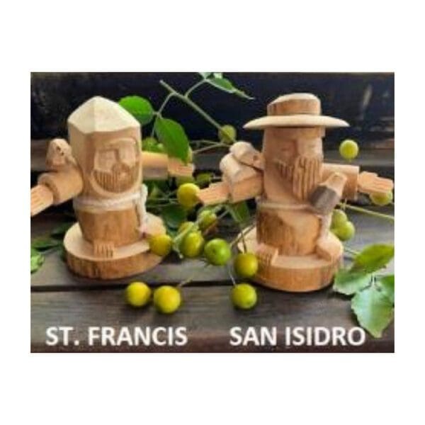 Two hand-carved wooden figurines, labeled St. Francis and the Hand Carved San Isidro Figure, are surrounded by green berries and leaves on a wooden surface.