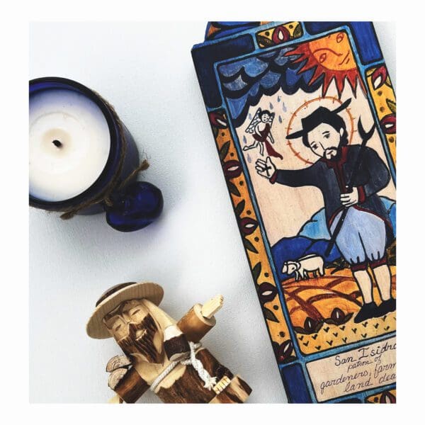 A SAN ISIDRO WOODEN RETABLO FOR FARMERS AND GARDENERS, featuring intricately carved and painted religious artwork of a man with farm animals, placed beside a small candle wrapped with a blue cord.