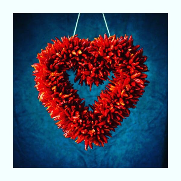 CHILE PEQUIN HEART WREATH, a red, heart-shaped decoration made of chili peppers, hanging against a blue background.
