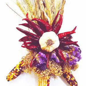 A decorative Chile Corn Ristra, measuring 7'' x 10'', featuring dried corn, red chillies, a garlic bulb, purple flowers, and wheat stalks.