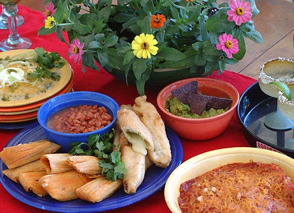 A table set with various Mexican dishes including tamales, stuffed peppers, beans, guacamole with blue corn chips, and a casserole. A potted plant with colorful flowers is in the background.